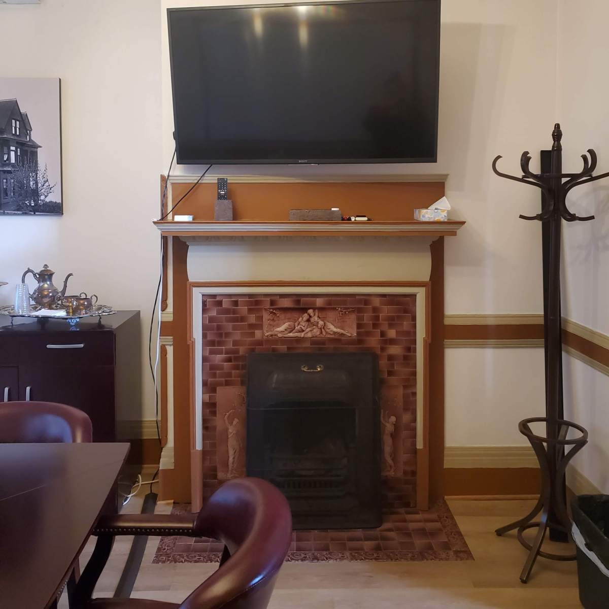 A living room with a mounted television above a fireplace, flanked by a coat rack and a dining table in the foreground.