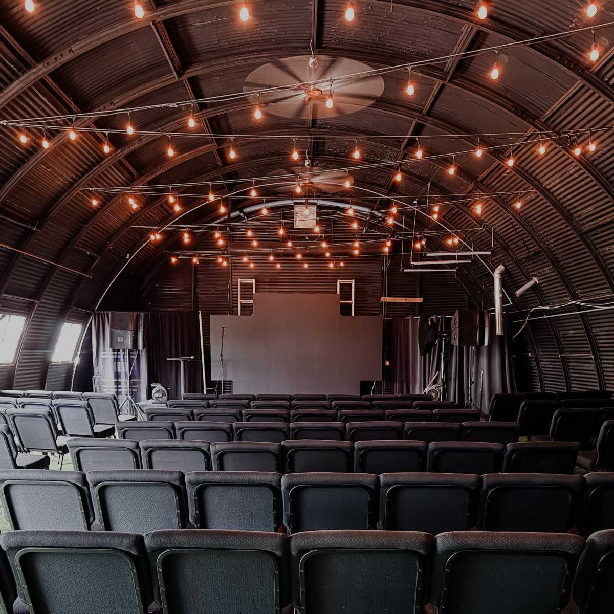 An interior view of a spacious event space featuring rows of black chairs facing a large screen, illuminated by string lights overhead.
