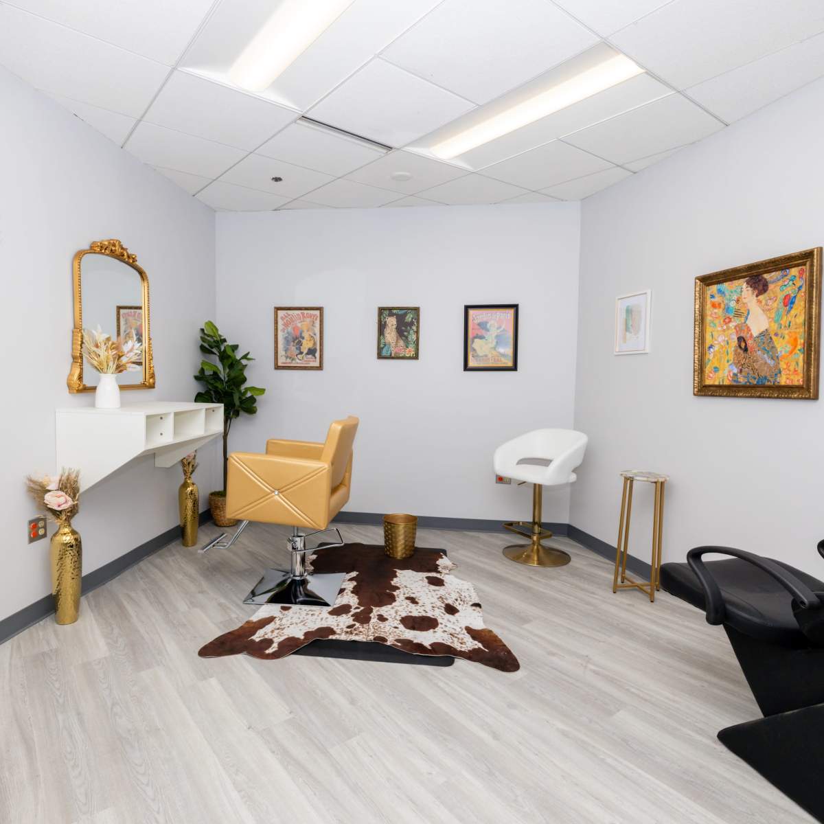A modern salon interior with a golden chair, a white vanity, framed artwork on the walls, and a cowhide rug on the floor.
