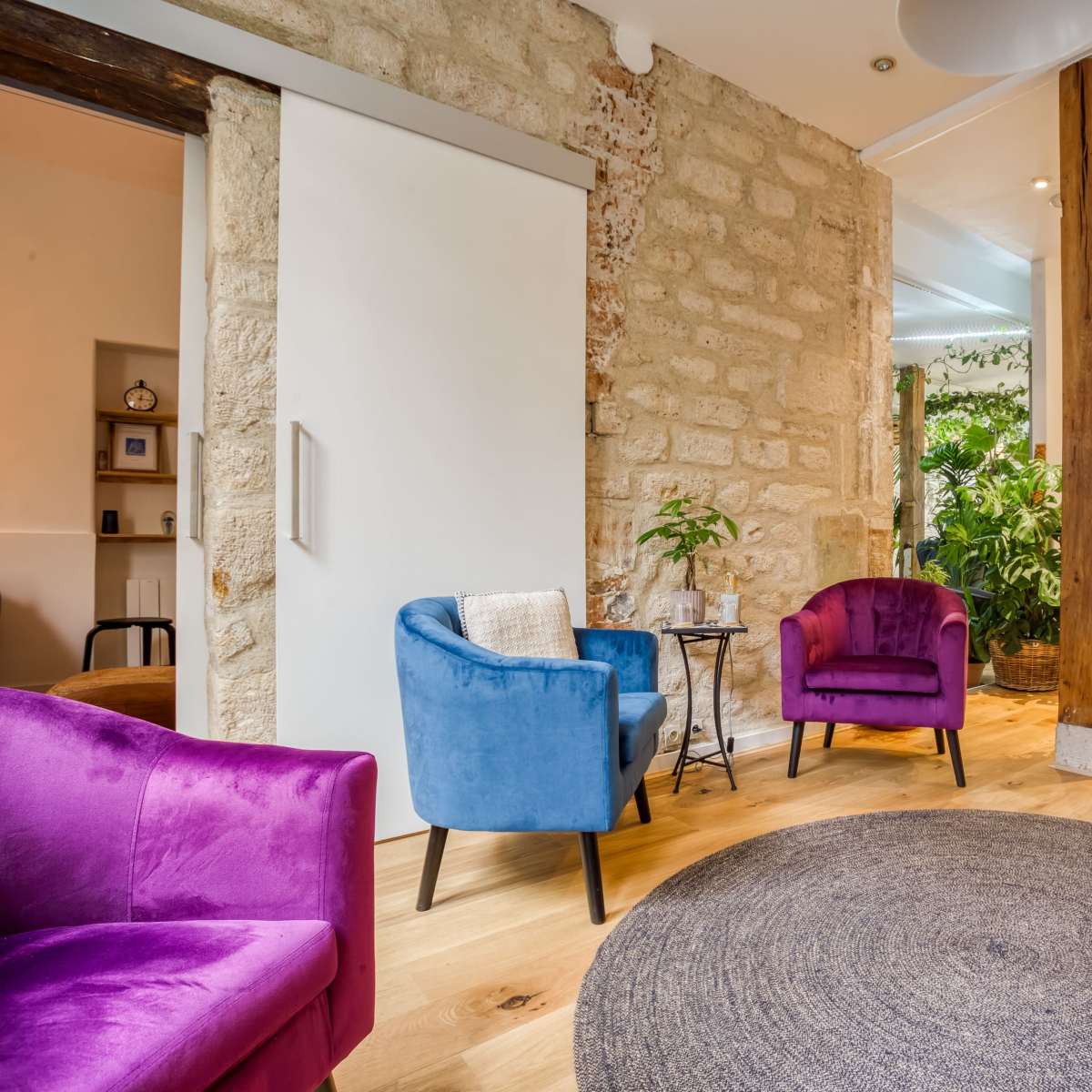 A modern interior space with two colorful chairs, a round rug, and wooden pillars surrounded by plants.