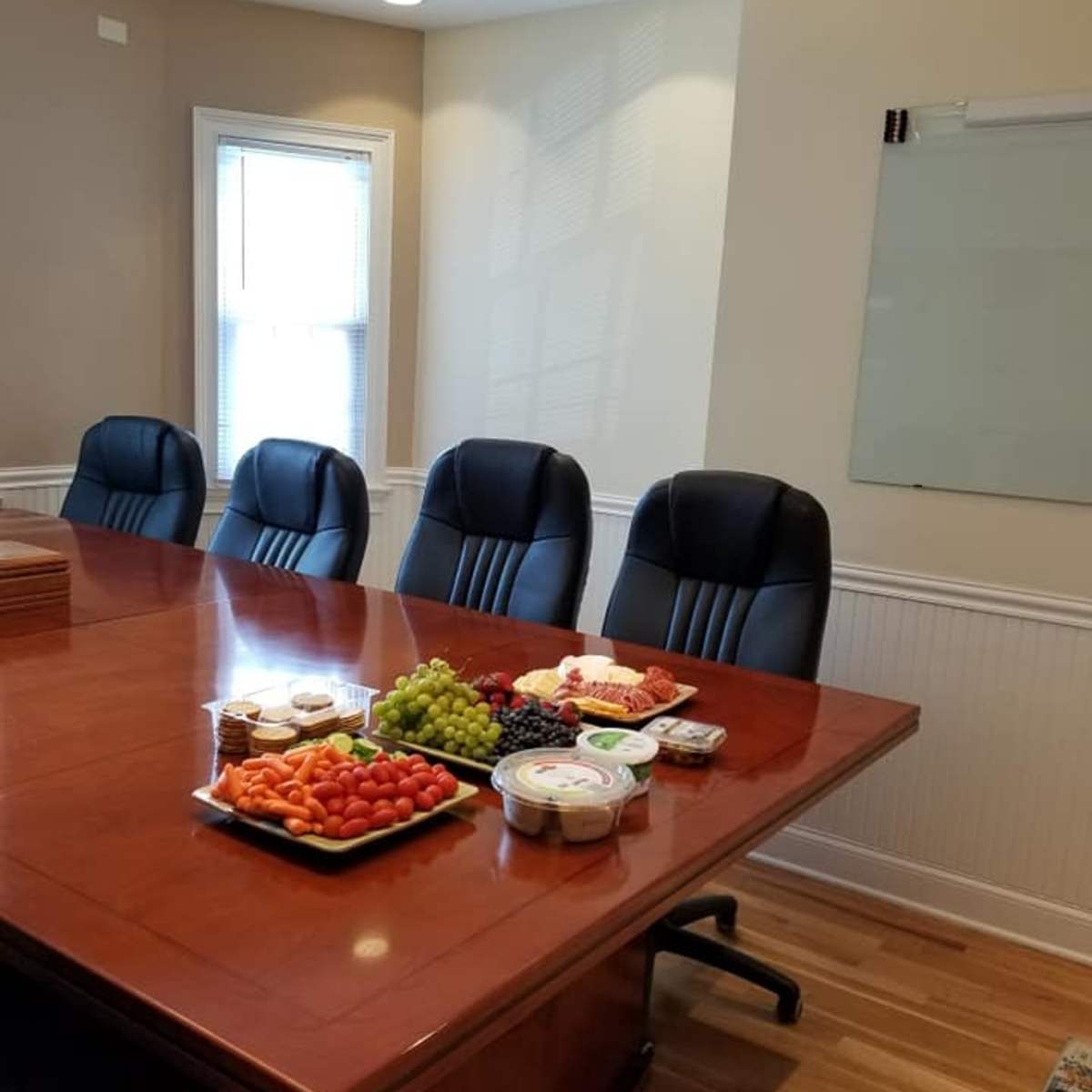A conference room is set up with a large wooden table displaying an assortment of fruits, vegetables, and snacks, while several black leather chairs are arranged around it.