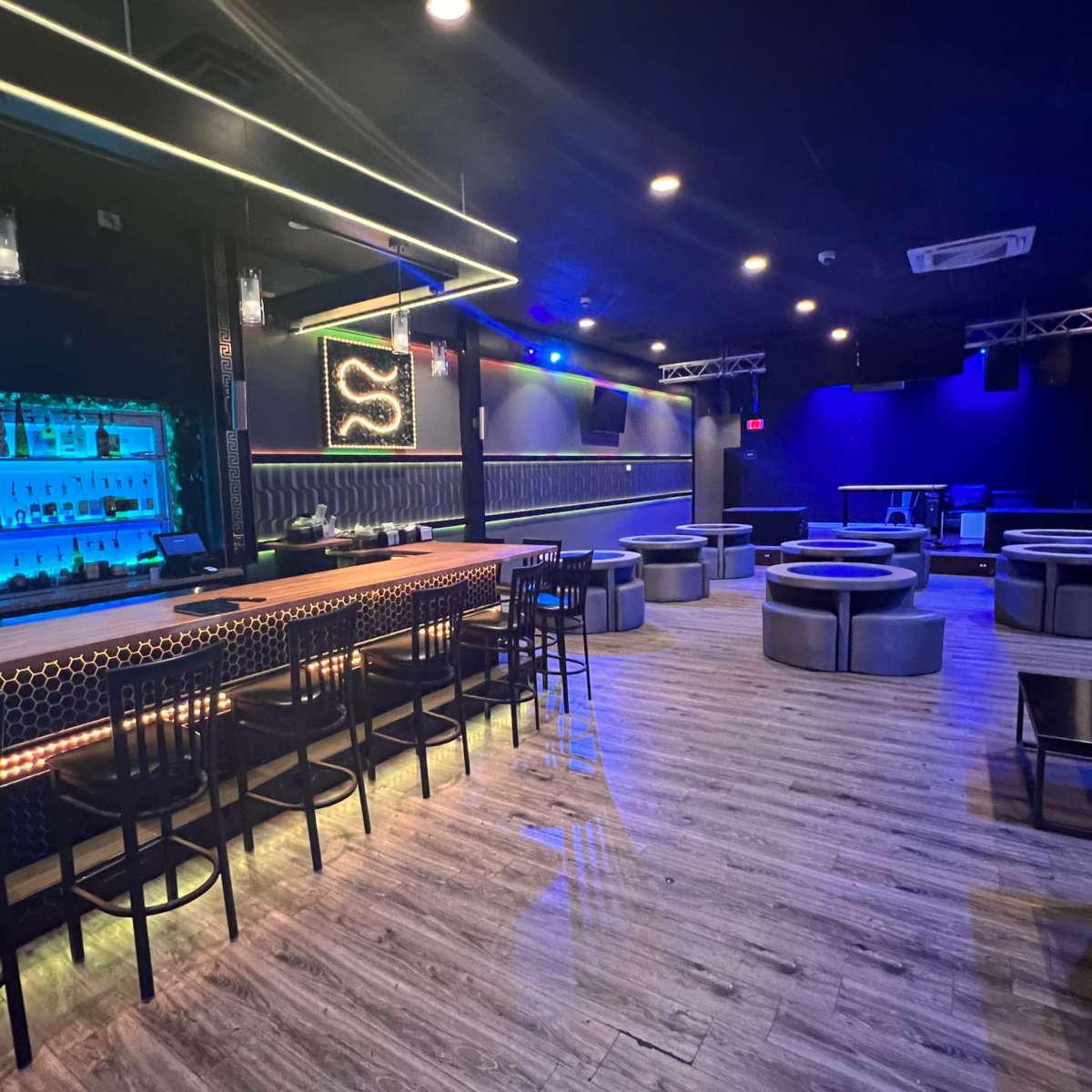A dimly lit bar area with a blue-lit backlit shelf displaying various bottles, surrounded by seating arrangements of circular booths and chairs.