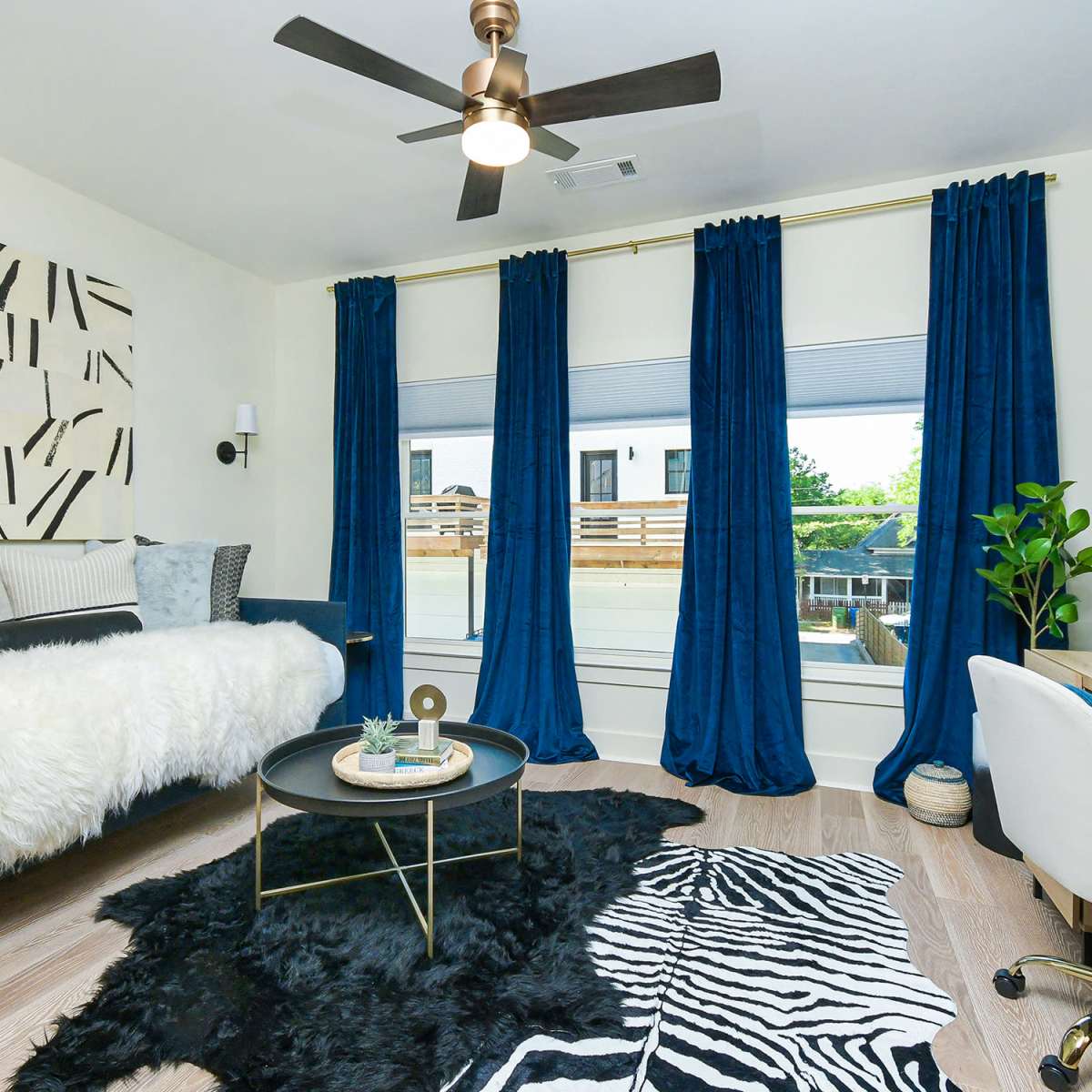 A modern bedroom features a daybed with a white faux fur throw, a black and white abstract artwork on the wall, dark blue curtains, a small desk, and a patterned rug.