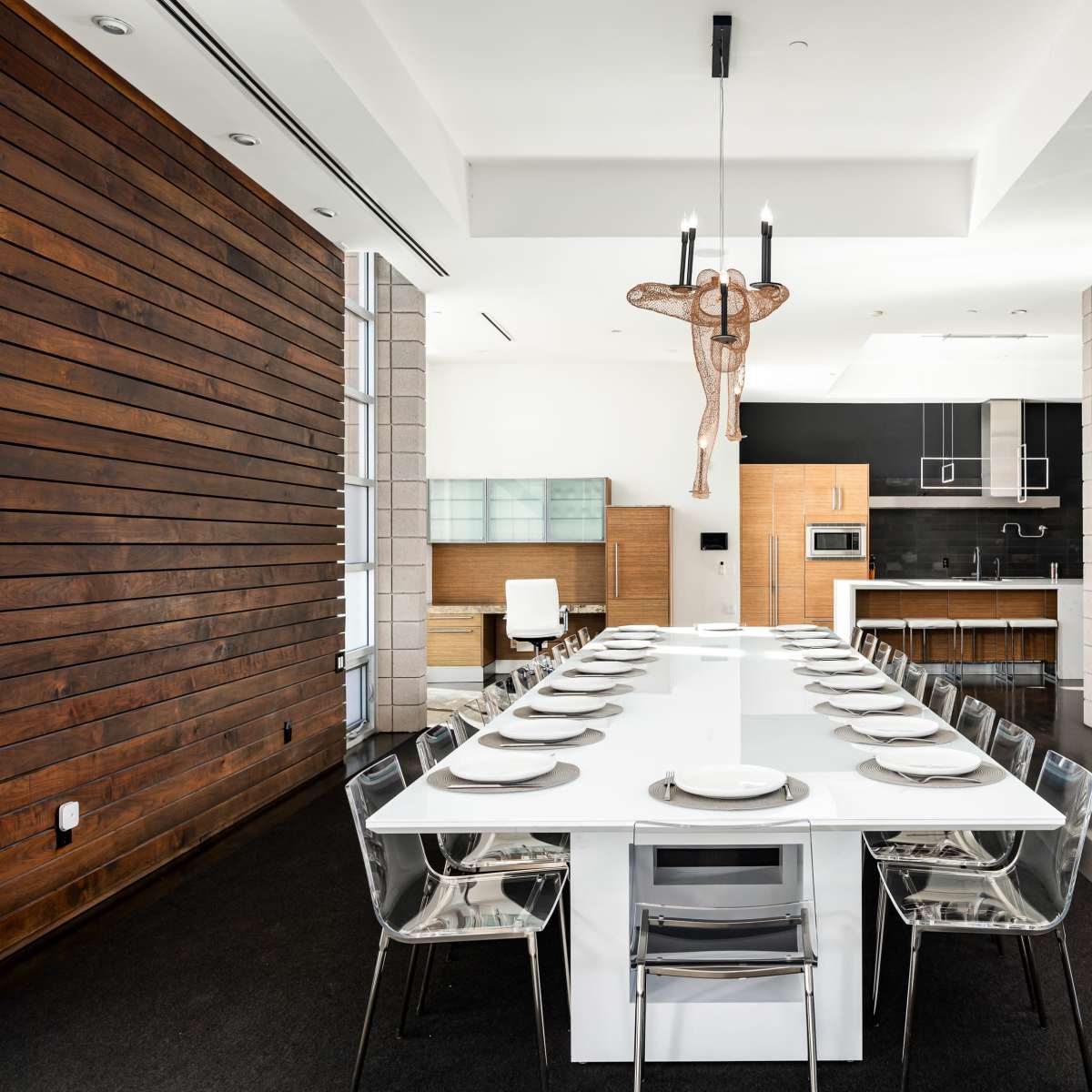 A modern dining area featuring a long white table surrounded by transparent chairs, with an open kitchen visible in the background.