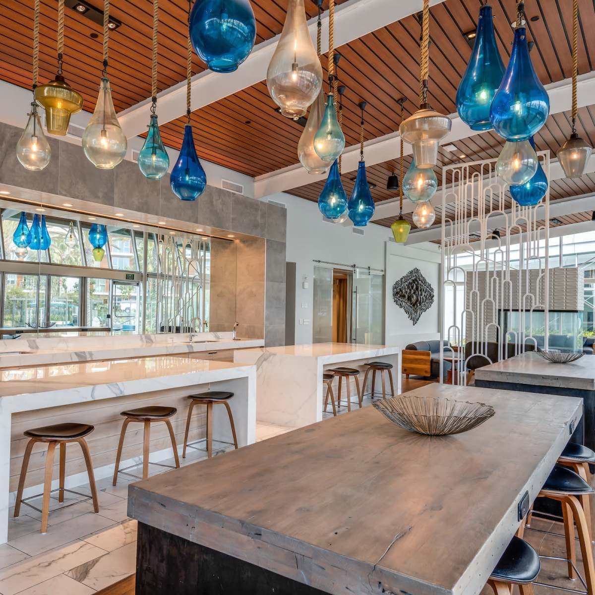 A modern kitchen with marble countertops, wooden bar stools, and colorful glass pendant lights hanging overhead.