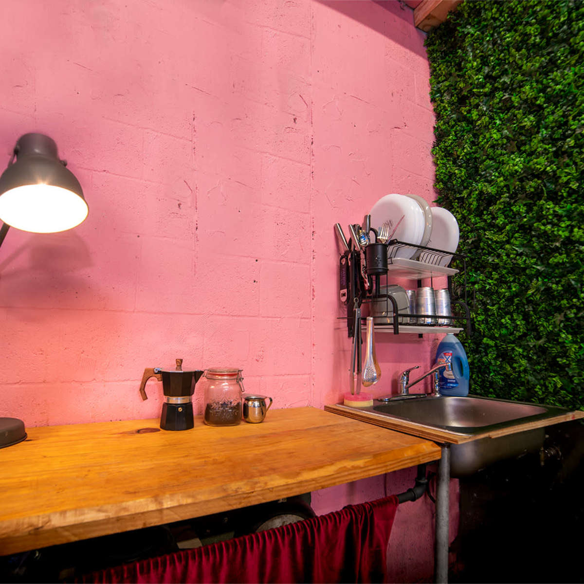 A kitchen corner with a pink wall, a wooden countertop, a sink, and shelves displaying dishes and kitchen tools, surrounded by artificial greenery.