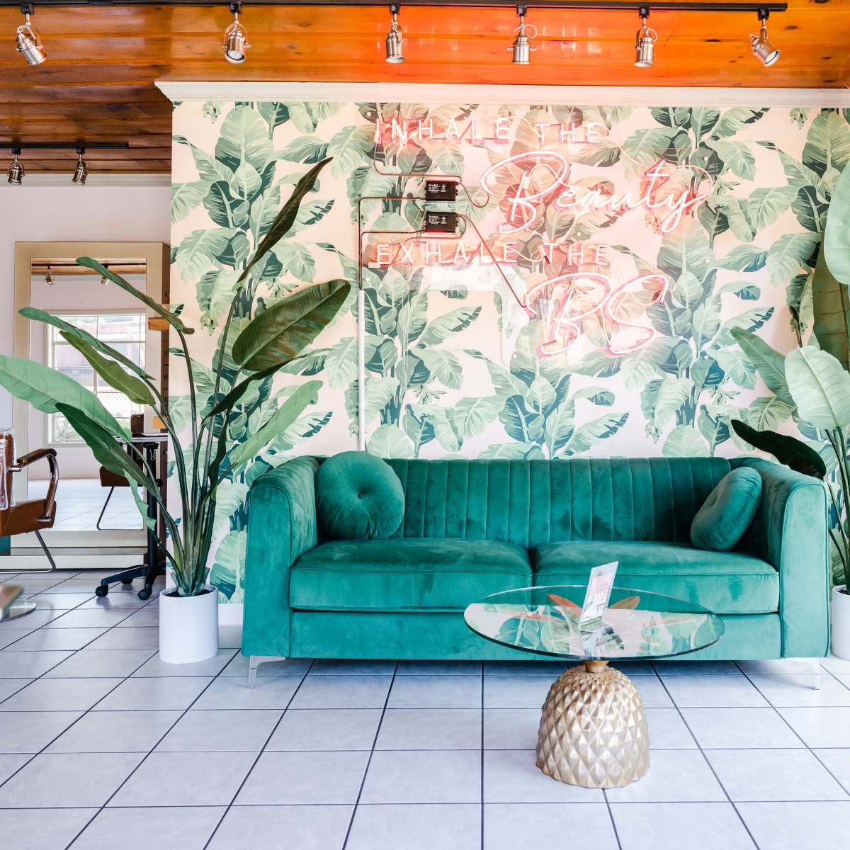 A room with a green velvet sofa in front of a leafy wallpaper, a glass coffee table, and large windows letting in natural light.