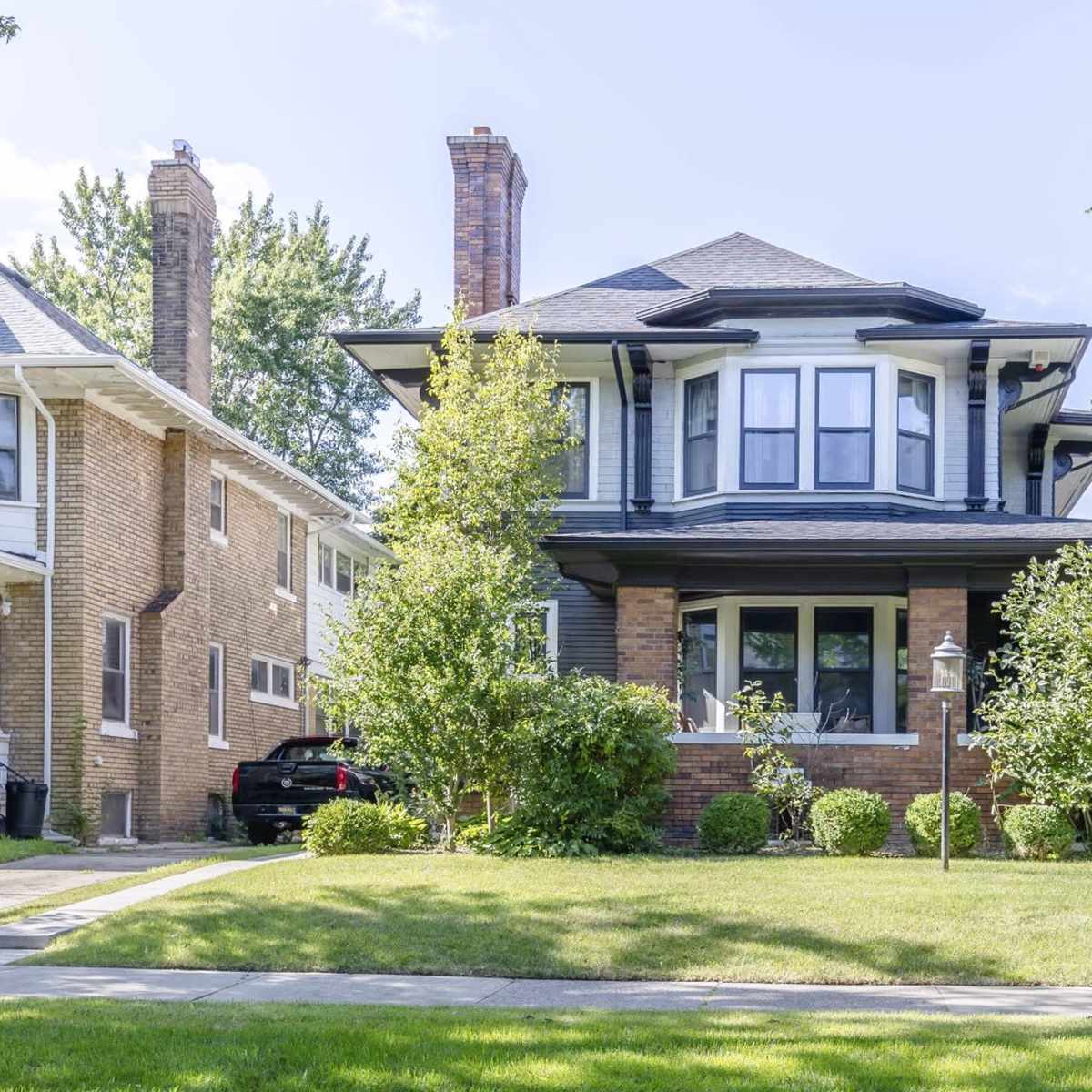 A two-story brick house with a wide porch and large bay windows stands surrounded by well-maintained shrubs and a lawn.