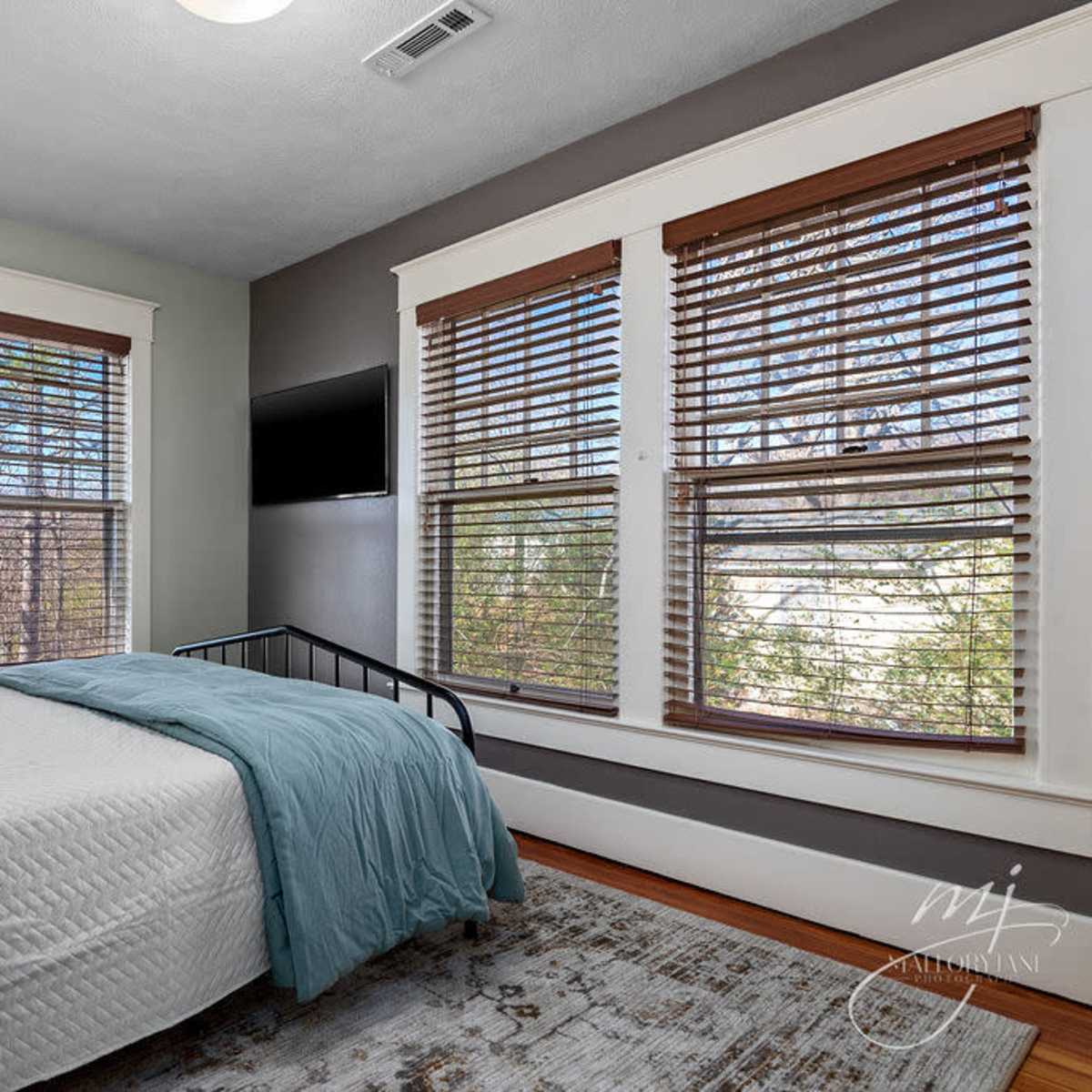 A bedroom features a bed with a light blue blanket, wooden blinds on the windows, and a flat-screen TV mounted on the wall.