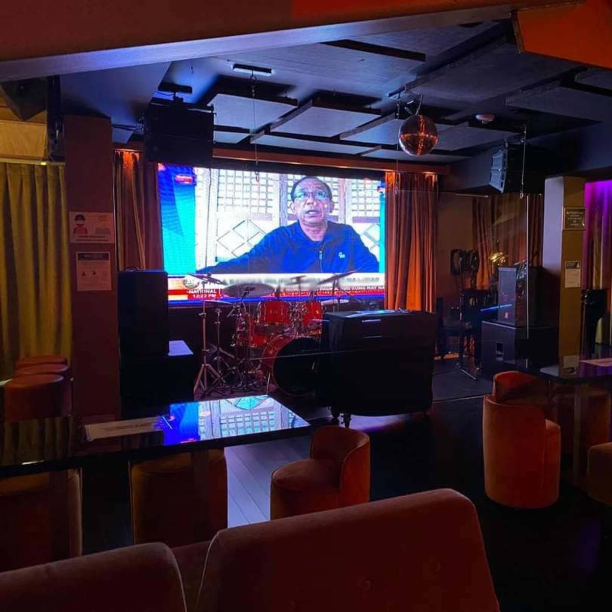 An empty lounge with a large screen displaying a news broadcast, a drum set, and seating arranged around dark tables.