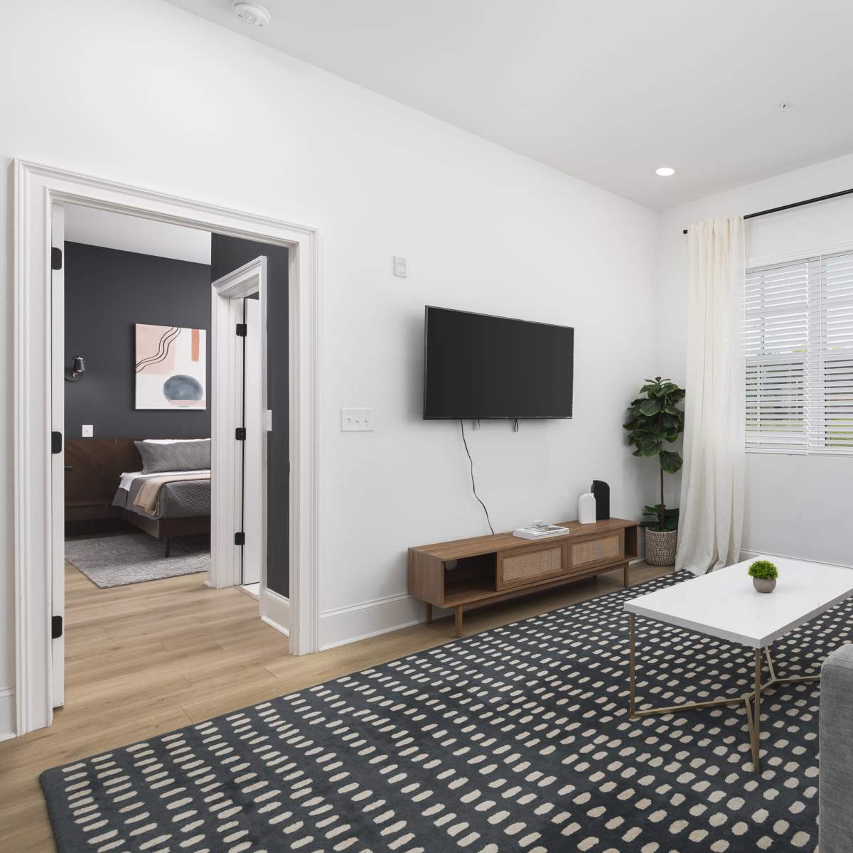 A modern living room features a gray sofa, a coffee table, and a television mounted on the wall, with a window dressed in curtains allowing natural light.