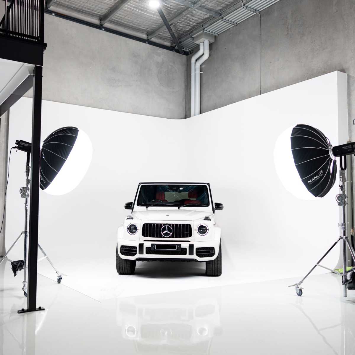 A white Mercedes-Benz G-Class is positioned in a well-lit studio with large softbox lights on either side and a seamless white backdrop.