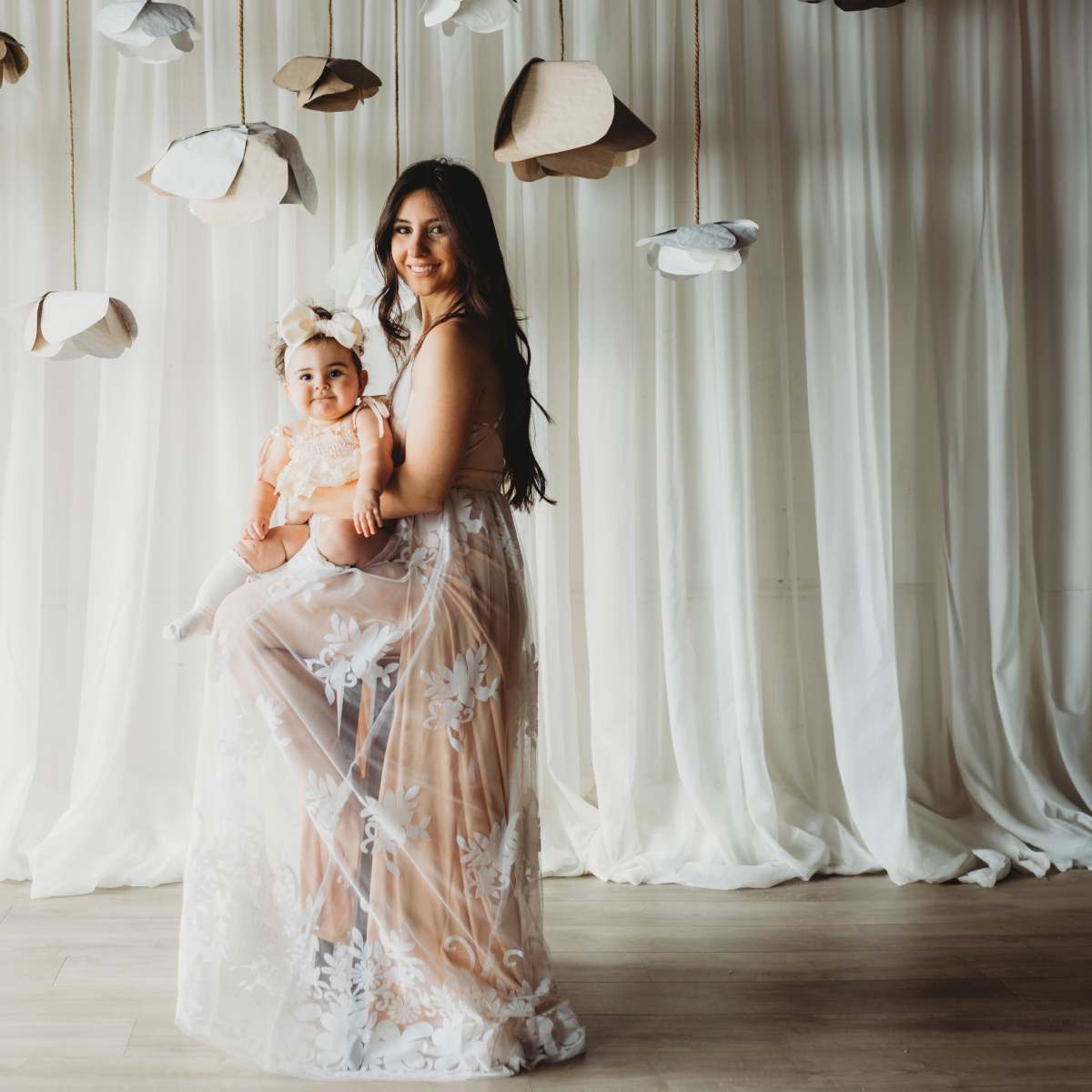 A woman holds a young girl as they stand against a backdrop of hanging paper clouds and draped fabric.