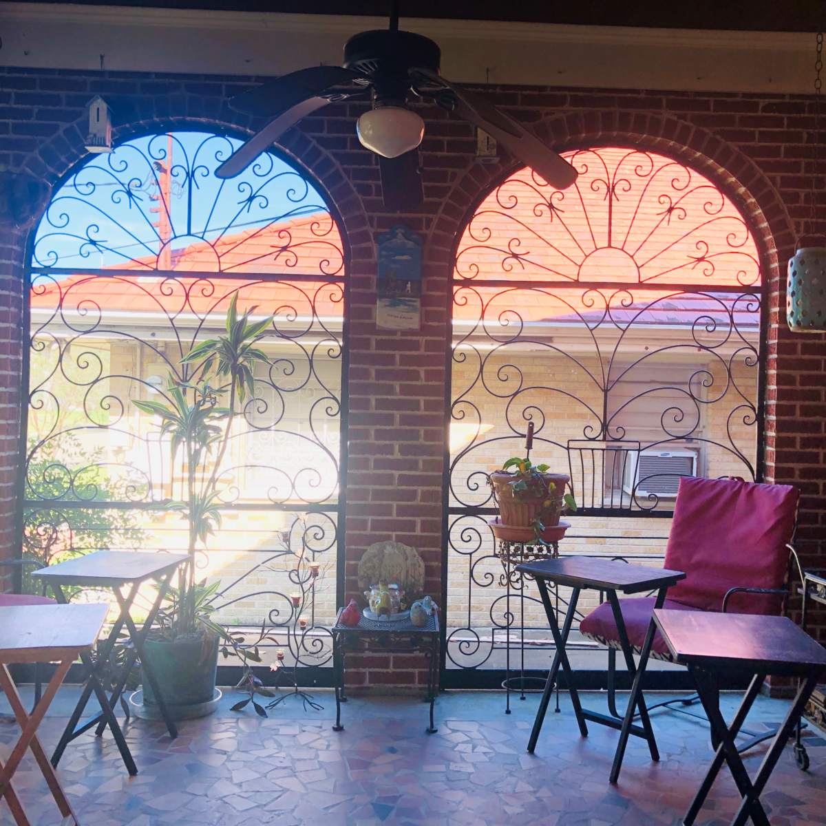 A brick porch with two arched openings, each featuring decorative wrought iron work, and wooden tables with chairs placed on a tiled floor.