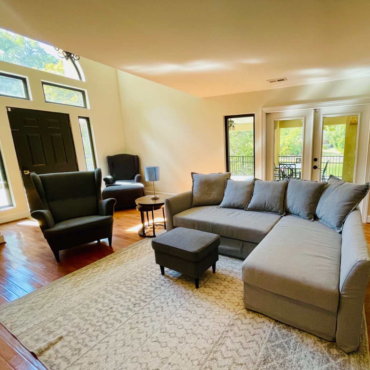 A cozy living room featuring a gray sectional couch, a small ottoman, a side table, and large windows overlooking a balcony.