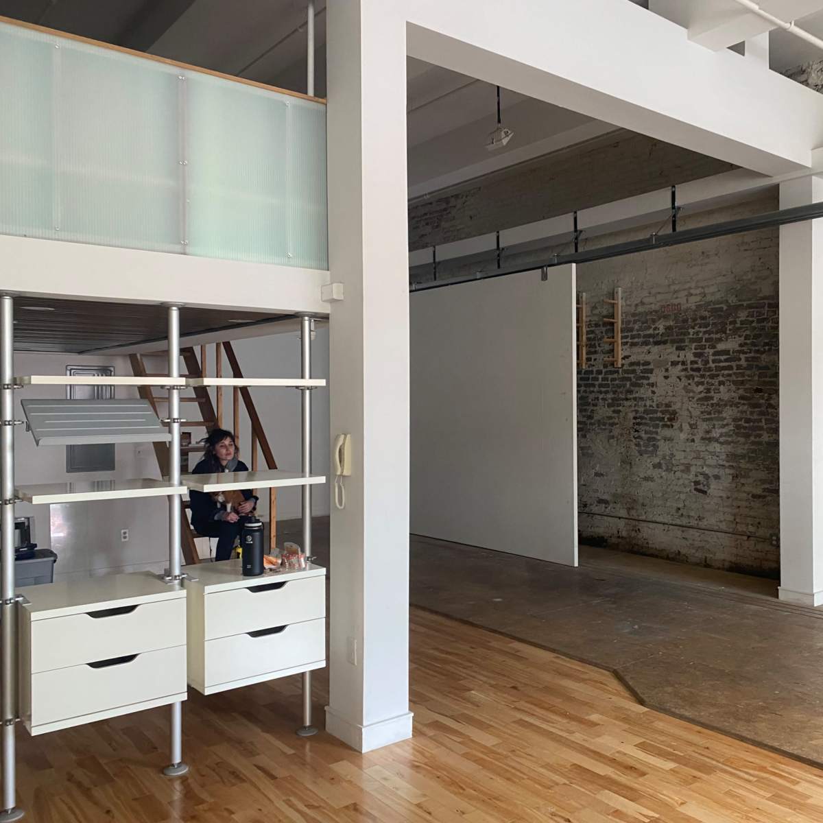 A spacious room with exposed brick walls, hardwood floors, and a minimalist office setup featuring a shelving unit and some furniture.
