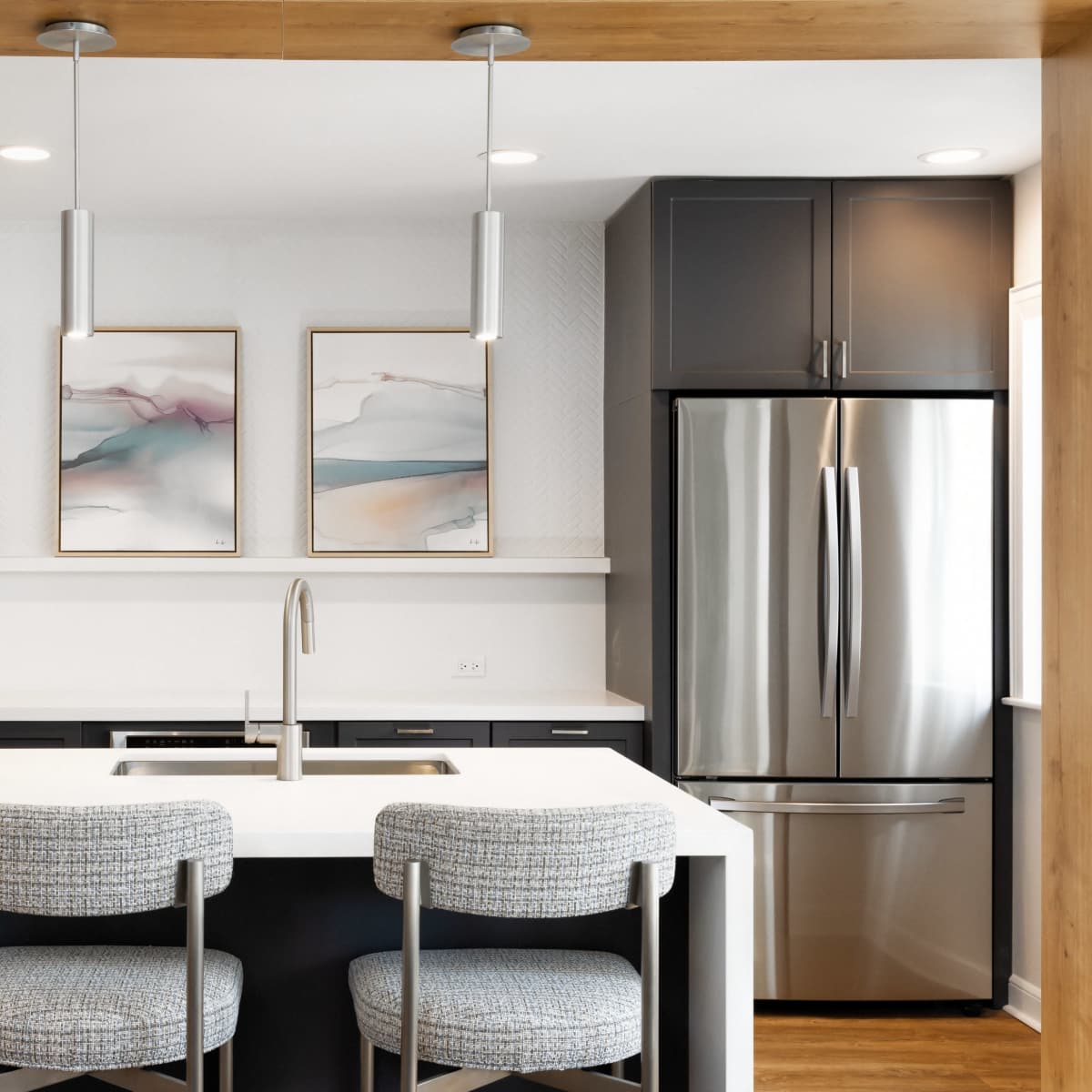 A modern kitchen features a central island with two bar stools, stainless steel appliances, and framed abstract art on the wall.