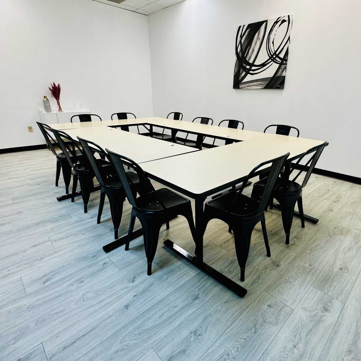 A conference room features a large rectangular table surrounded by black chairs, set against a minimalist white wall with abstract art.