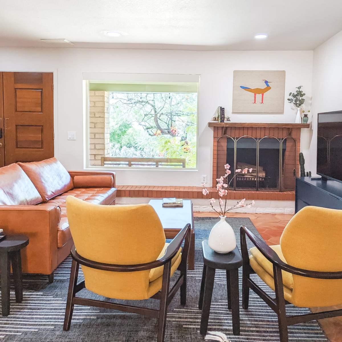 The living room features a brown leather sofa, two yellow chairs, a coffee table, and a television facing a decorative fireplace beneath a window.