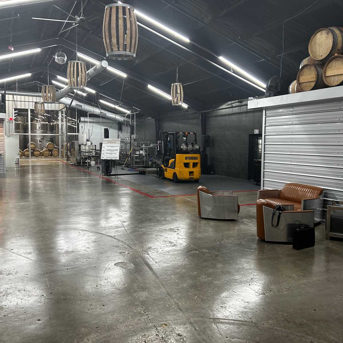 An industrial space with wooden barrels, a forklift, and various seating arrangements, illuminated by overhead lights.