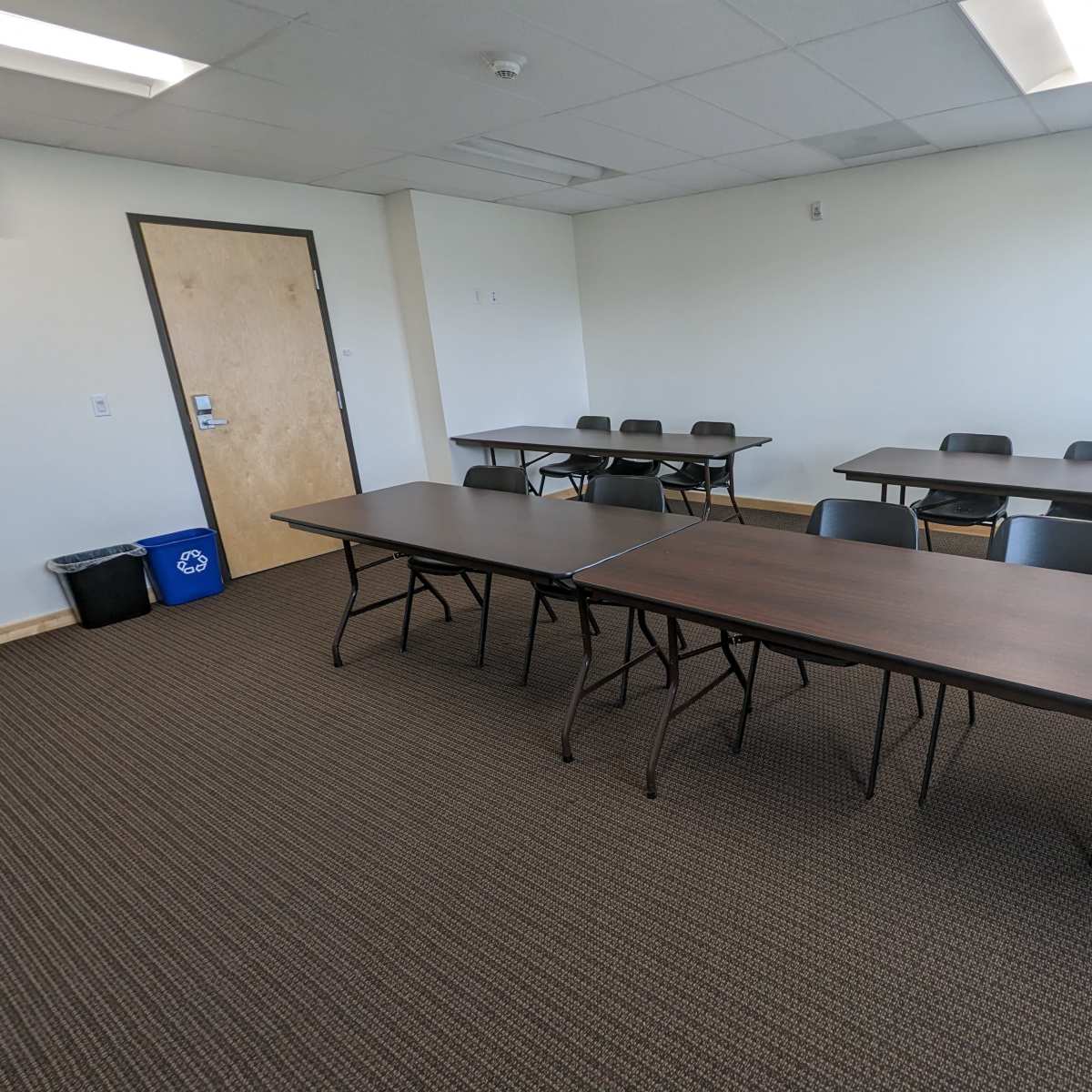 A meeting room features two rows of rectangular tables and chairs with a door and recycling bin in the corner.