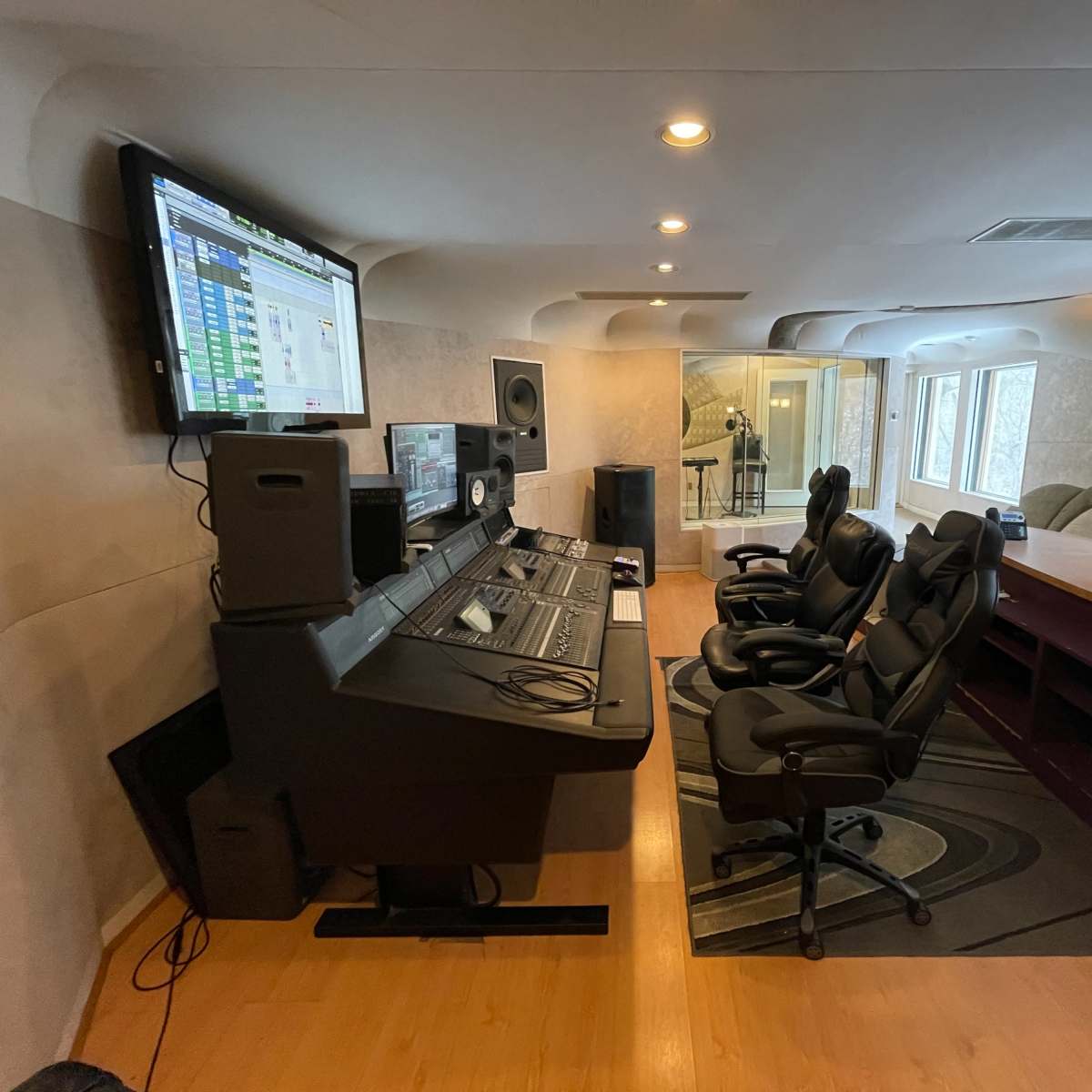 A modern recording studio with a large mixing console, computer monitors, and several ergonomic chairs arranged in front of a control desk.