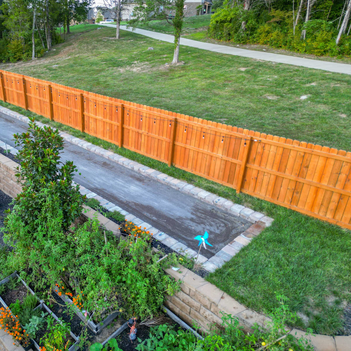 A fenced backyard featuring a concrete pathway and garden beds with various plants along the edges.