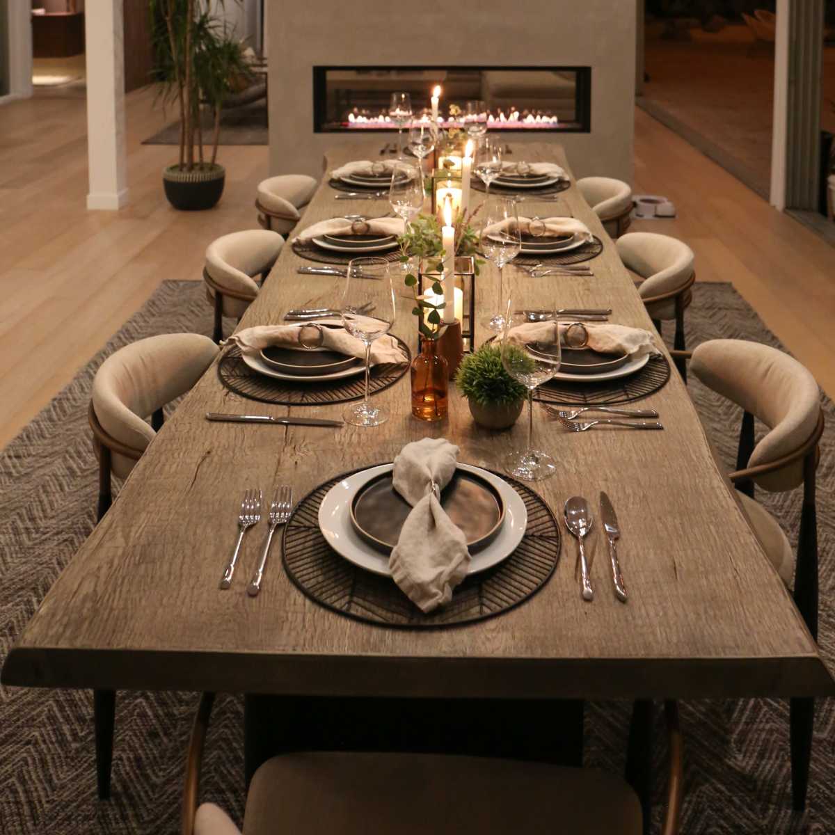 A long dining table is set with plates and silverware, featuring candles and plants, beside a modern fireplace in an open-concept space.