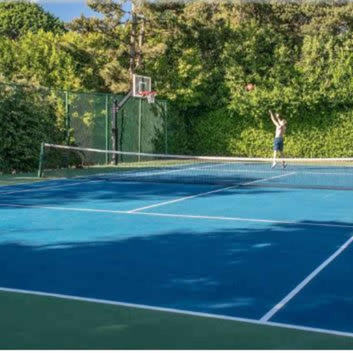 A person is shooting a basketball on a court adjacent to a tennis court surrounded by greenery.