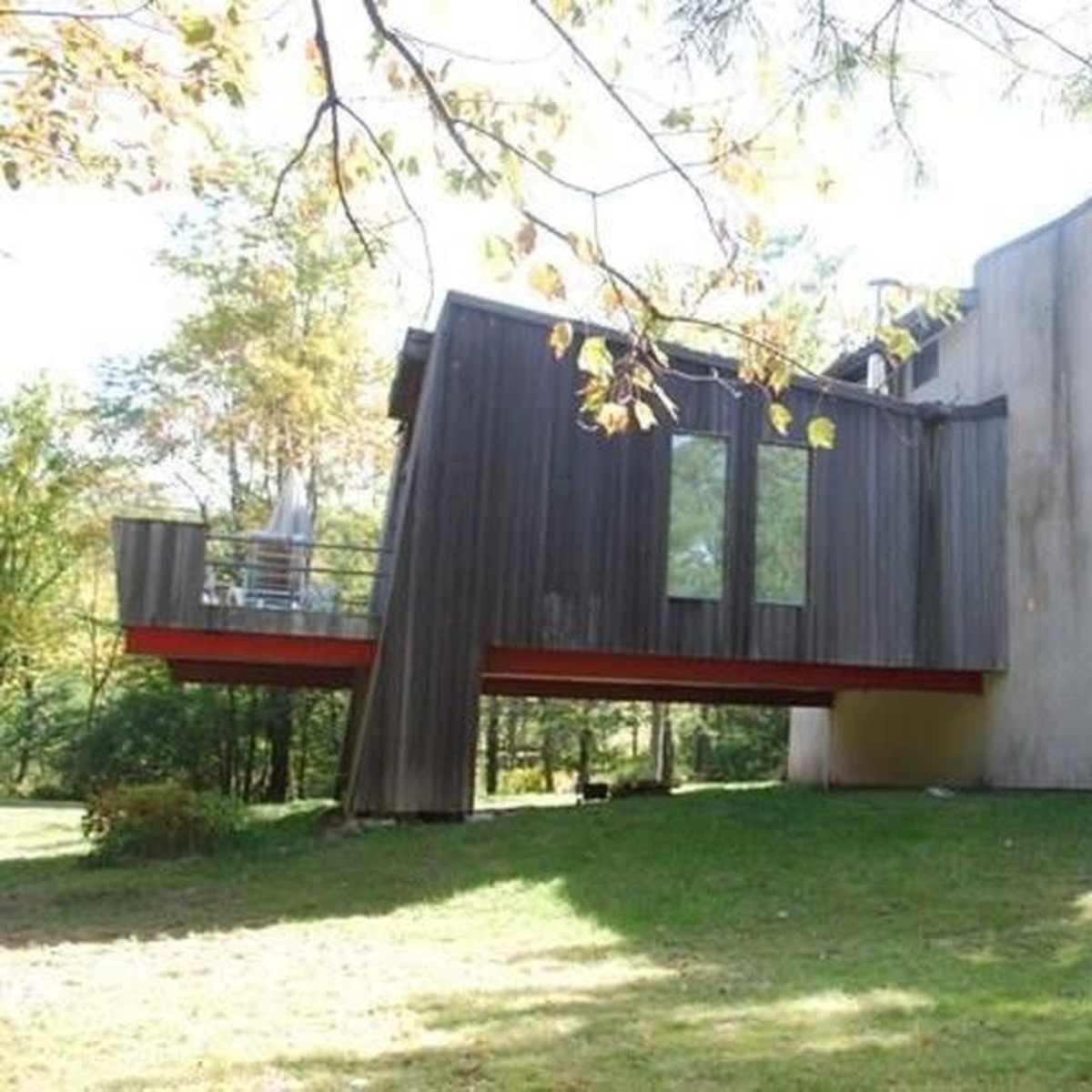 A modern house with a combination of wooden and concrete materials, positioned on a grassy area surrounded by trees.