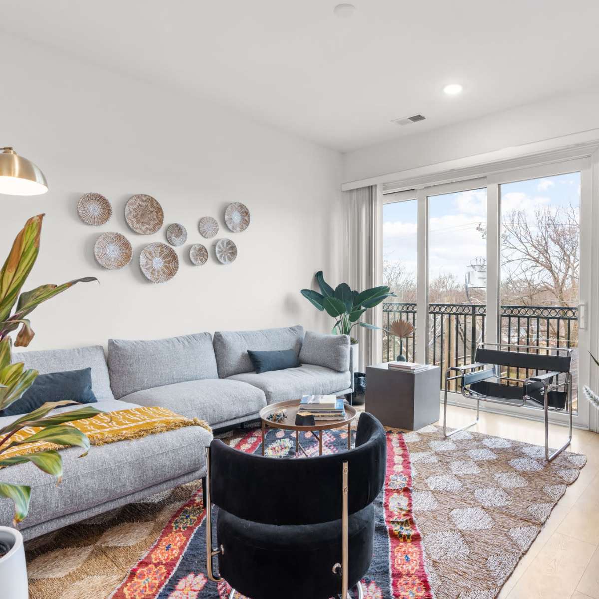 A modern living room features a gray sectional sofa, a round coffee table, and a TV against a white wall with various decorative pieces, complemented by large windows that open to a balcony.