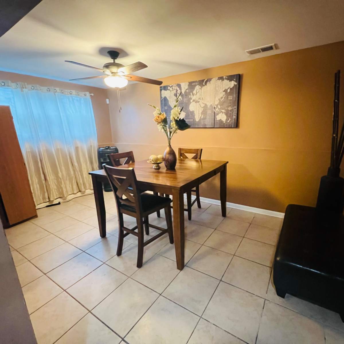 A dining area features a wooden table with chairs, a vase of flowers, a world map on the wall, and a partially visible curtain window.