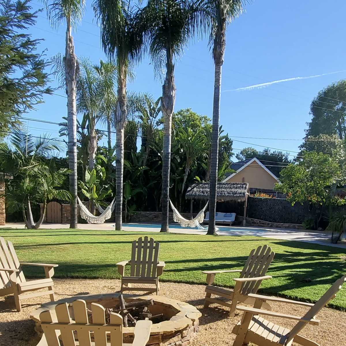 A backyard features a fire pit surrounded by several white adirondack chairs, with palm trees and a swimming pool in the background.