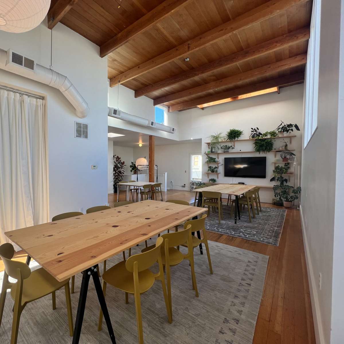 A spacious room features wooden beams, multiple tables with chairs, and greenery, with large windows allowing natural light.