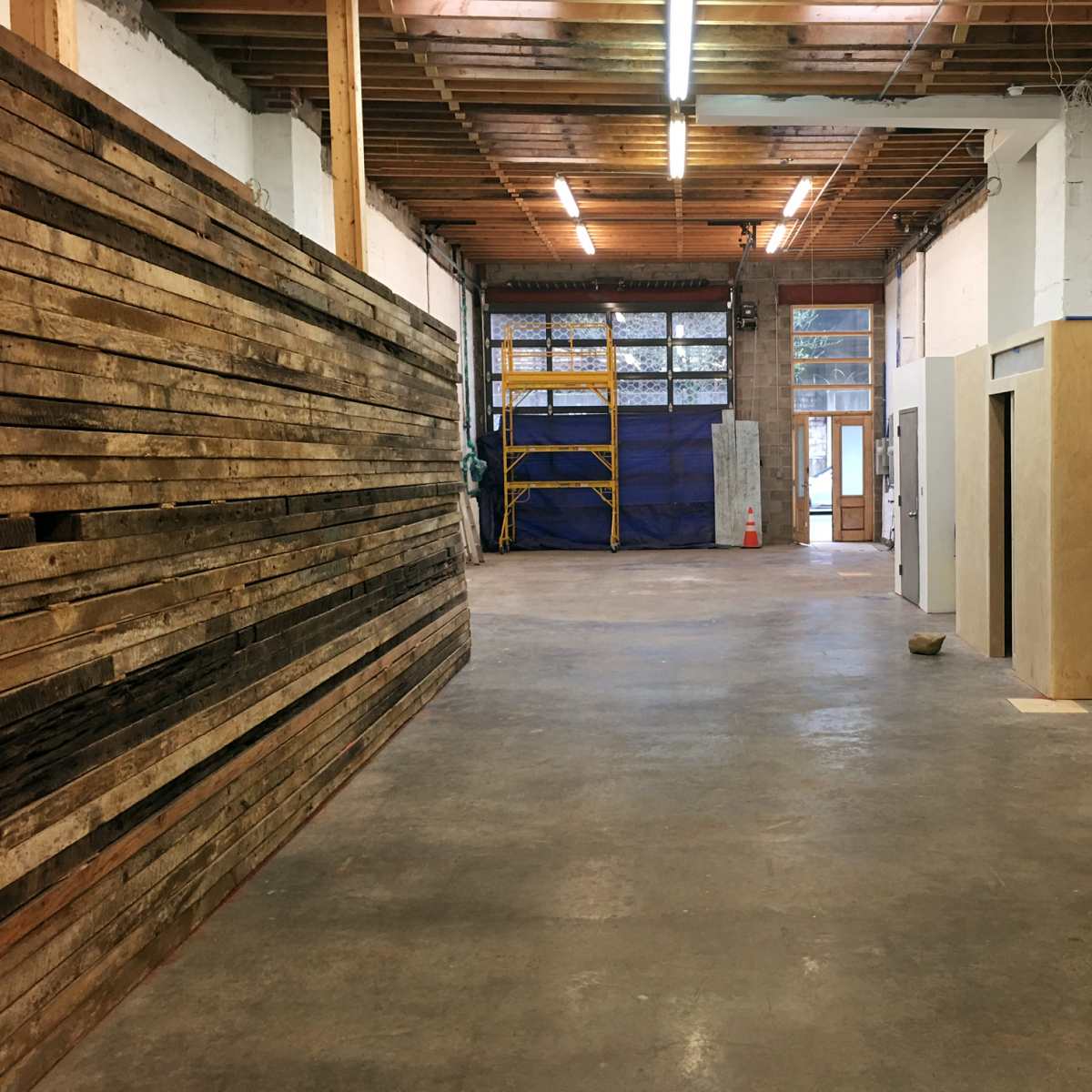 A spacious interior of a workshop or construction area with wooden walls on one side, an unfinished wall space, and scaffolding in the background.