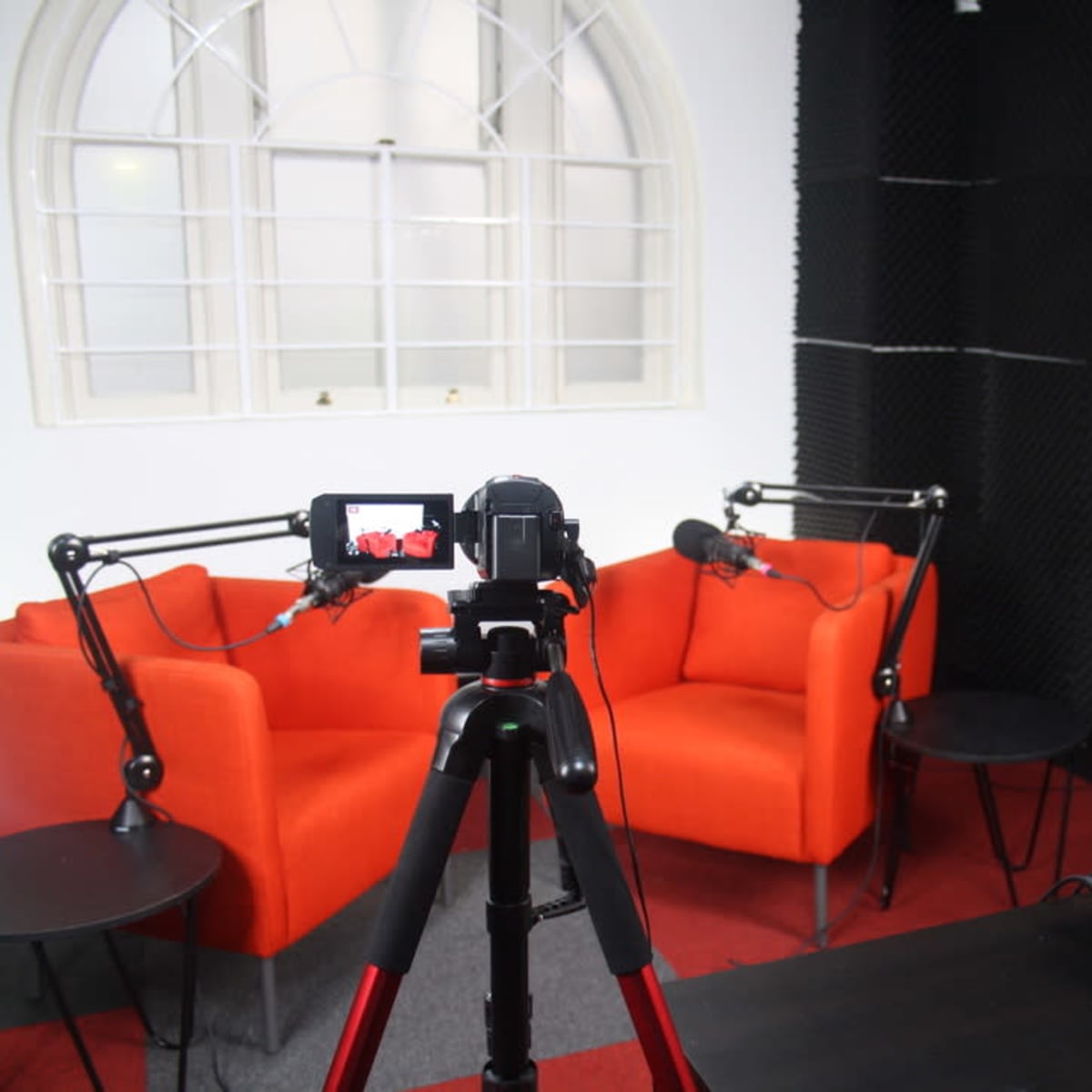 A recording studio with two orange chairs, a microphone setup, and a video camera on a tripod in front of a control panel.