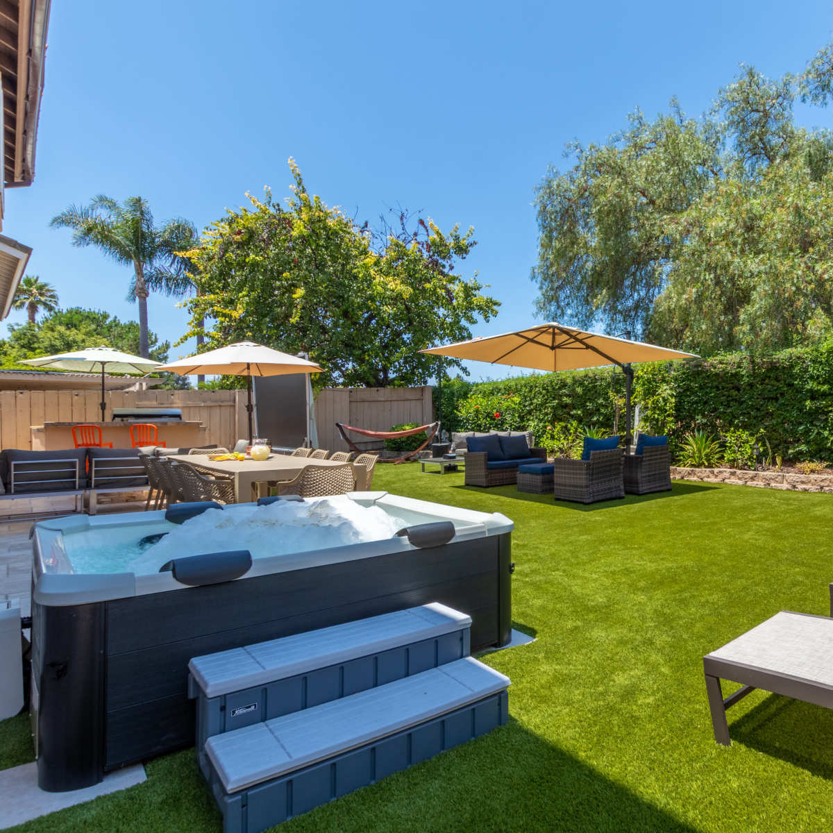 A landscaped backyard with a hot tub, lounge chairs, umbrellas, and a dining area surrounded by greenery.