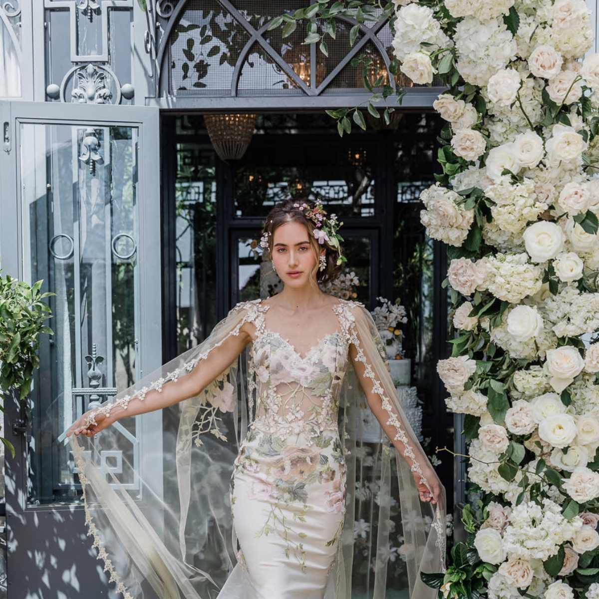 A bride in an intricate wedding gown with floral embellishments walks through a doorway adorned with greenery and white flowers.