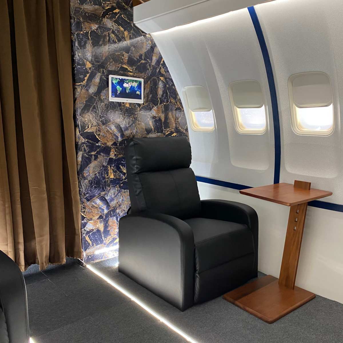 A sleek black armchair next to a wooden side table, positioned in front of a section of an airplane interior with multiple windows and a textured wall.