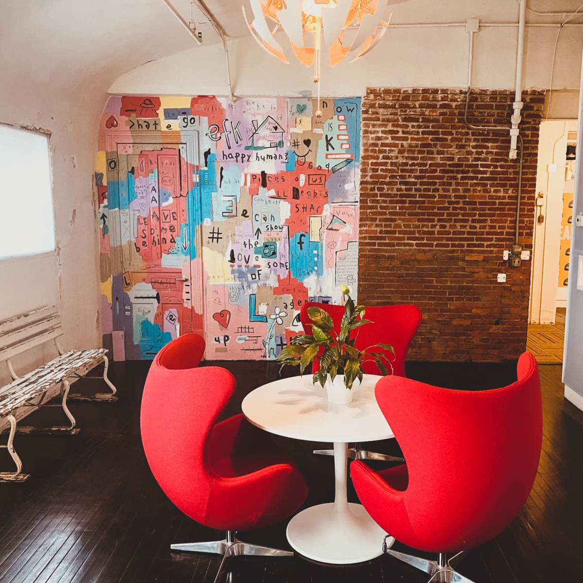 A small dining area features a round white table surrounded by three red chairs, with a colorful mural on the wall and a plant at the center of the table.