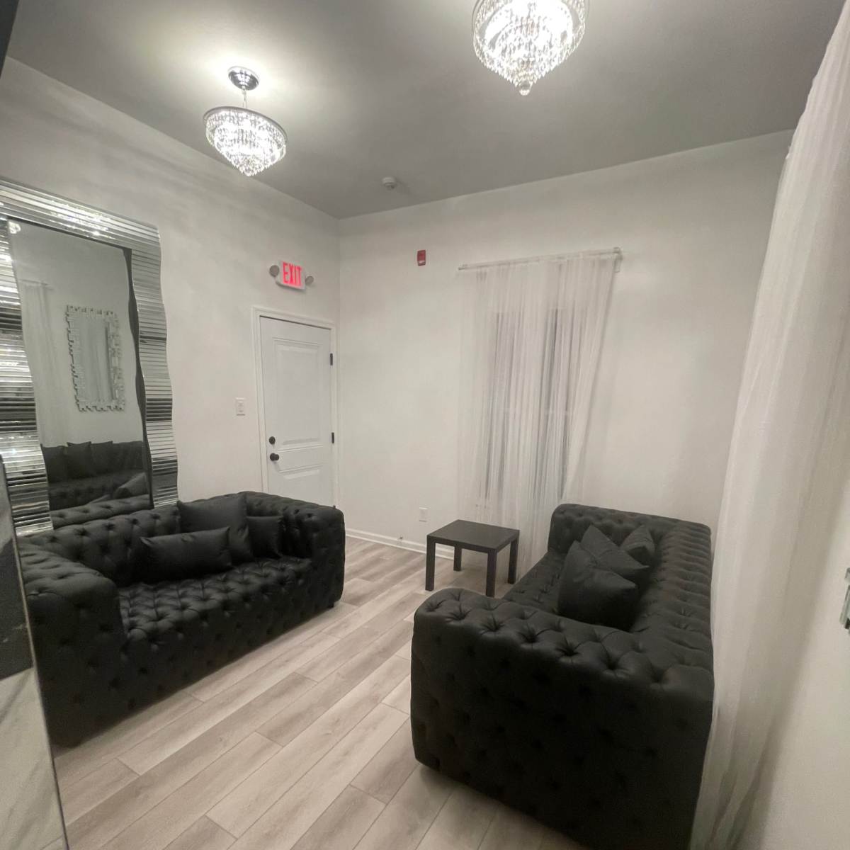 A minimalistic room with two black tufted sofas facing a small table, accented by two crystal chandeliers and a mirror on one wall.