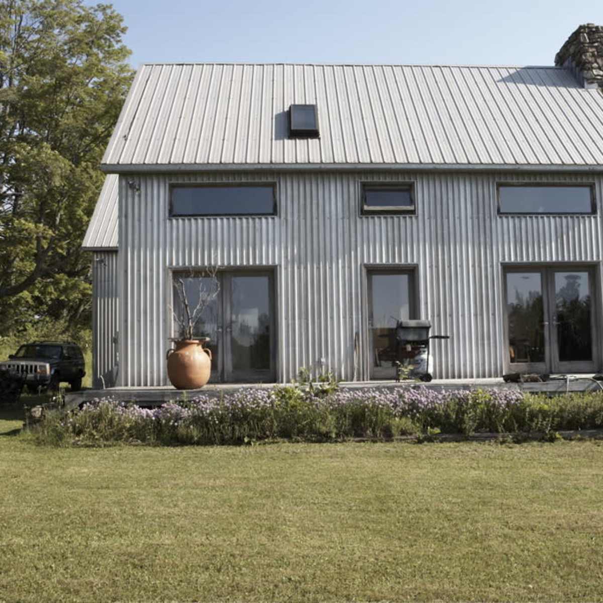 A modern metal-clad house with large windows is situated on a lush green lawn surrounded by trees and a flower bed.