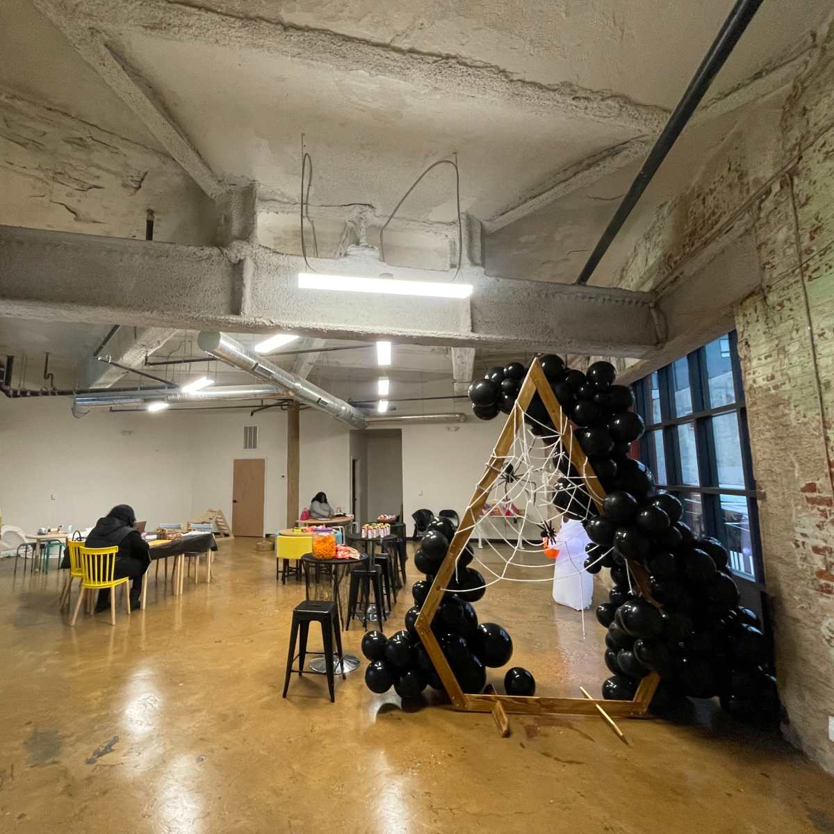 An industrial-style room with exposed brick walls, a large window, and a black balloon arch decorated with spider web, while several people are engaged in activities at tables.