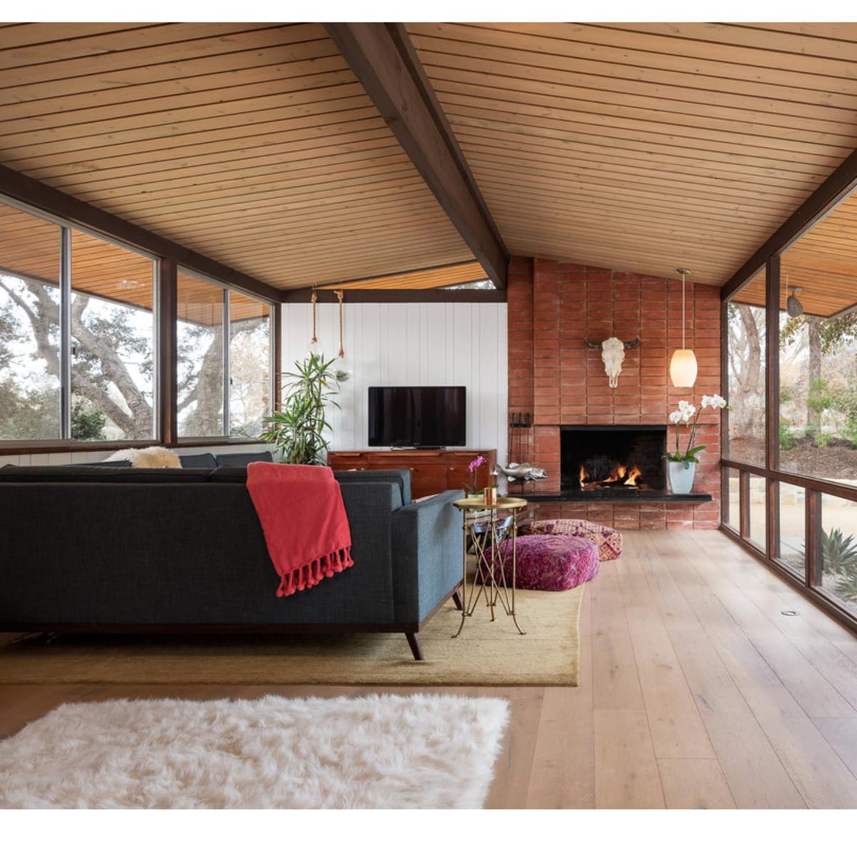 A modern living room with large windows, wooden ceilings, and a fireplace surrounded by a carpet, furniture, and decorative plants.