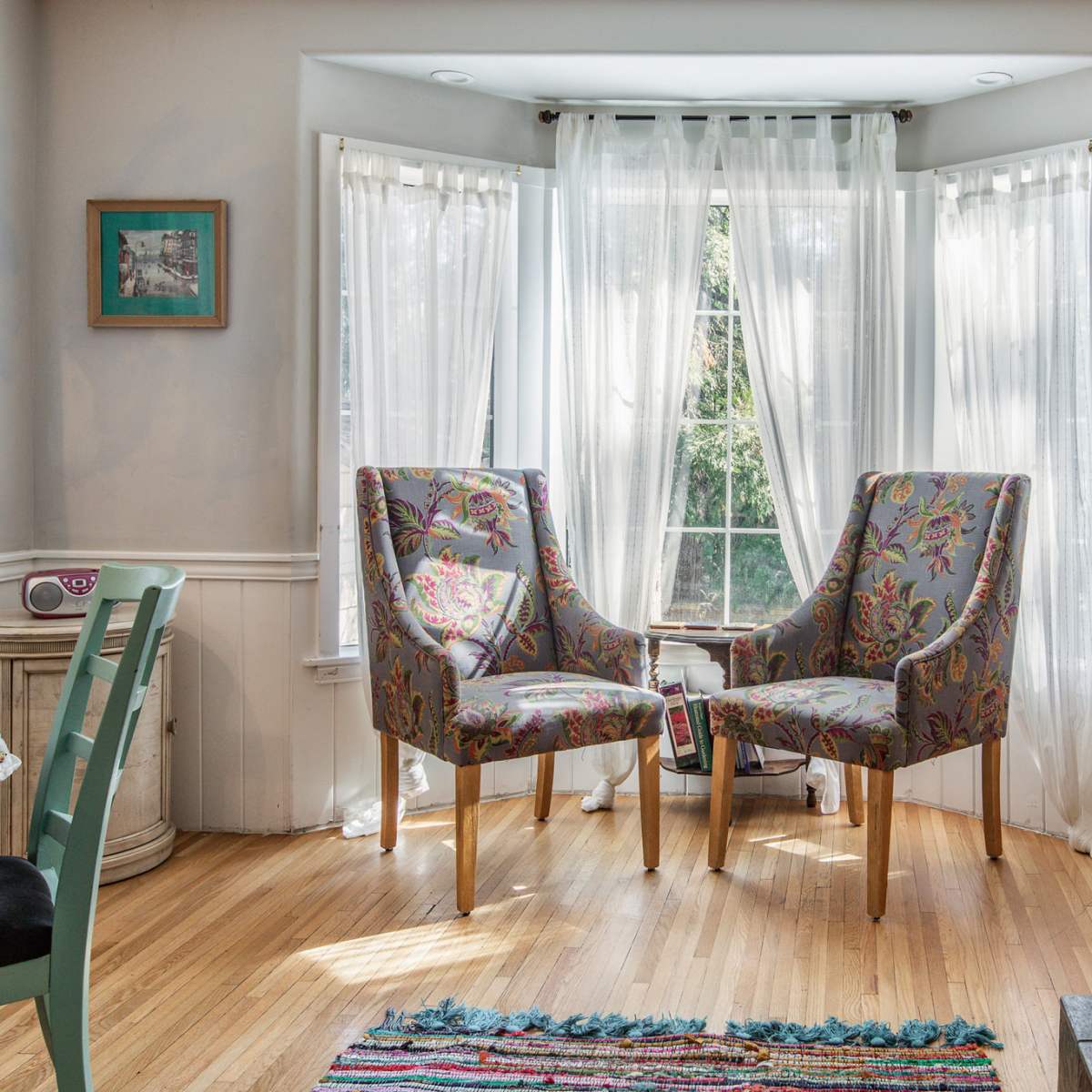 A bright corner of a room features two floral-patterned chairs facing each other near a large window, with light curtains allowing natural light to filter in.