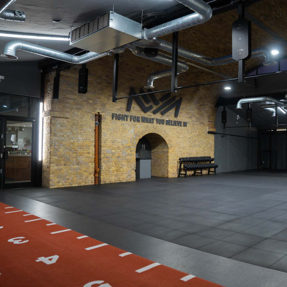 A spacious gym with a brick archway, a black rubber floor, and motivational text on the wall.
