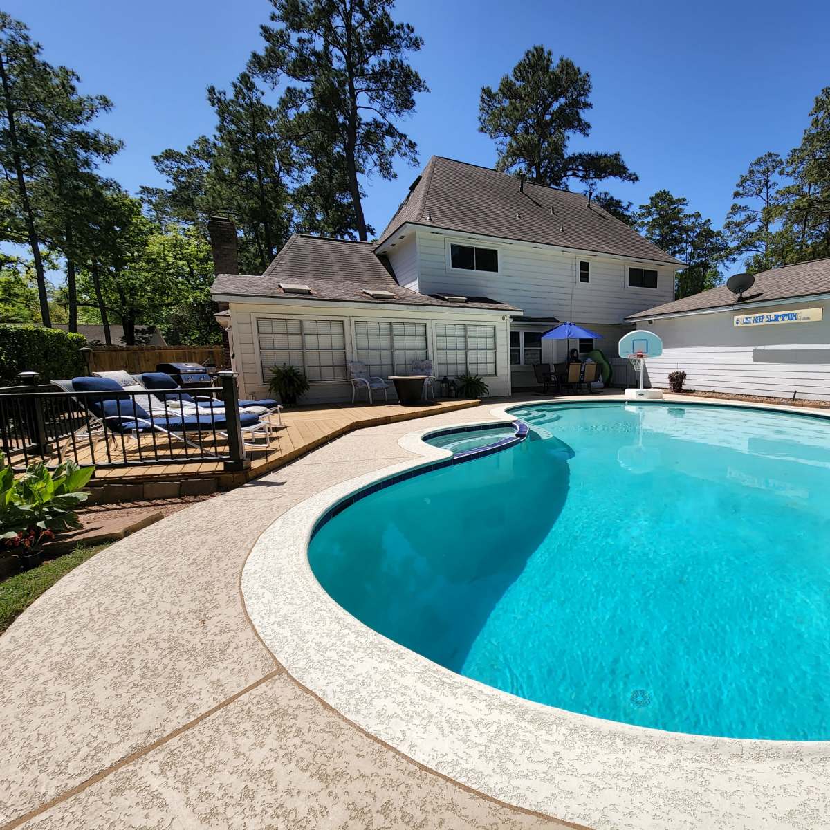 A clear blue swimming pool surrounded by a patio, lounge chairs, and a house with a basketball hoop in a wooded backyard setting.
