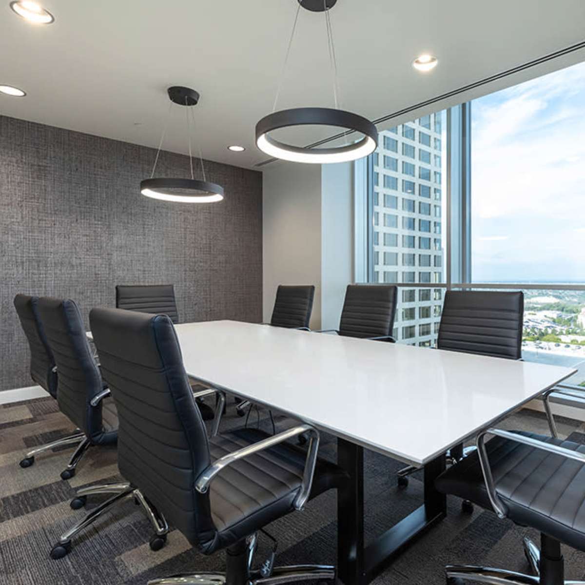 A modern conference room features a white rectangular table surrounded by eight black cushioned chairs, with large windows providing a view of the city skyline.