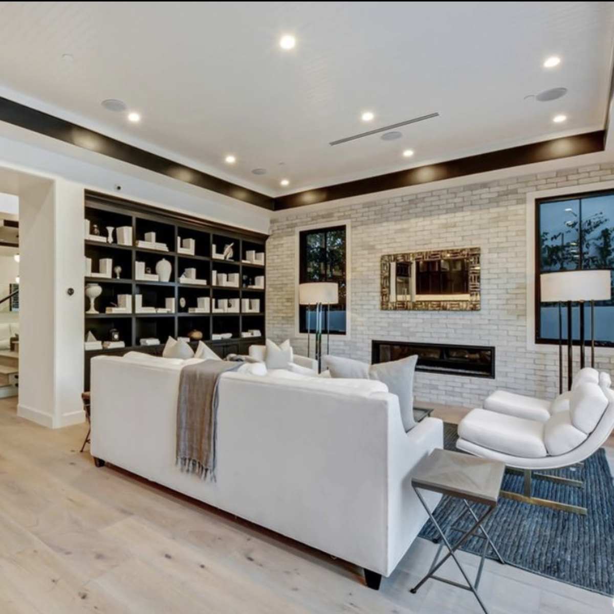 A modern living room with white furniture, a fireplace, and large windows, featuring a minimalist bookshelf and light wood flooring.