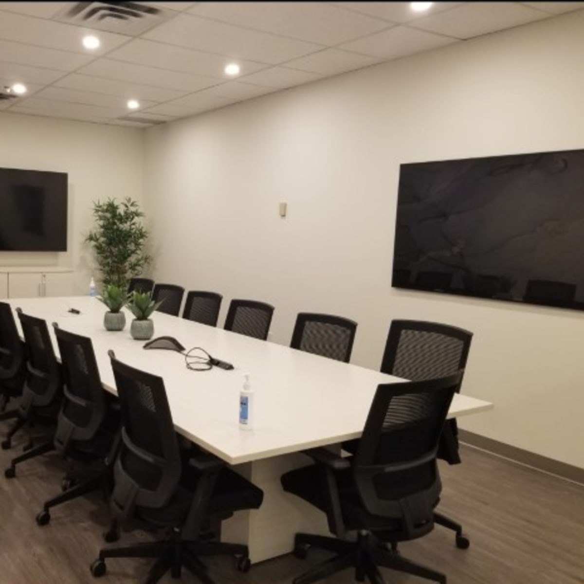 A modern conference room features a long table surrounded by ergonomic chairs, with two large screens mounted on the walls and potted plants in the corners.